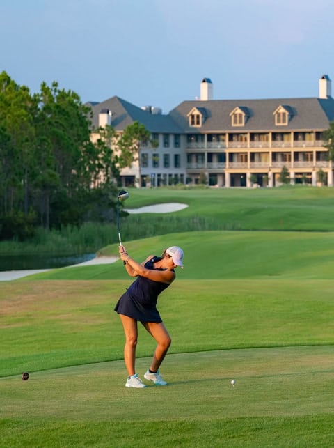 A woman in a navy dress swings a golf club on a golf course with a large building and trees in the background.